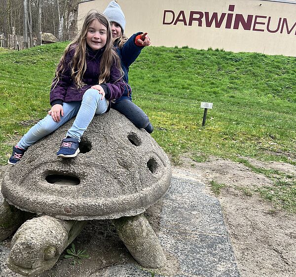 Zwei Mädchen sitzen auf einer steinernen Schildkröte. Im Hintergrund das Darwineum.