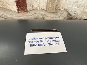 Auf einer dunklen Spendentruhe steht ein weißes Schild "Biblia Nova Pauperum. Spende für die Fenster. Bitte helfen Sie uns."
