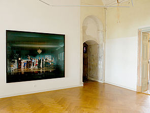 Ein querformatiges Gemälde füllt die Raumwand. Darauf stehen in einem dunkelgrünen Raum Menschen wie bei einem Familienbild nebeneinander
