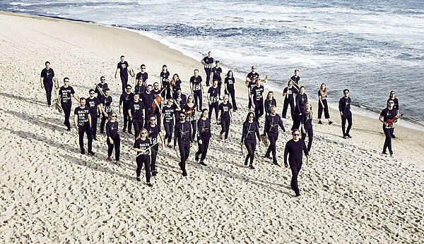 Das Baltic Sea Philharmonic und Dirigent Kristjan Järvi gehen am Strand entlang. Im Hintergrund liegt die Ostsee. Eine Luftaufnahme.