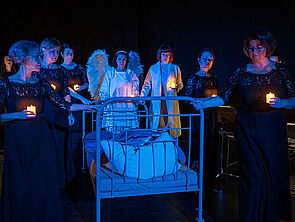 Auf einer dunklen Bühne stehen mehrere Frauen. Jede hält eine Kerze in der Hand. In der Mitte steht ein weißes Gitterbett.