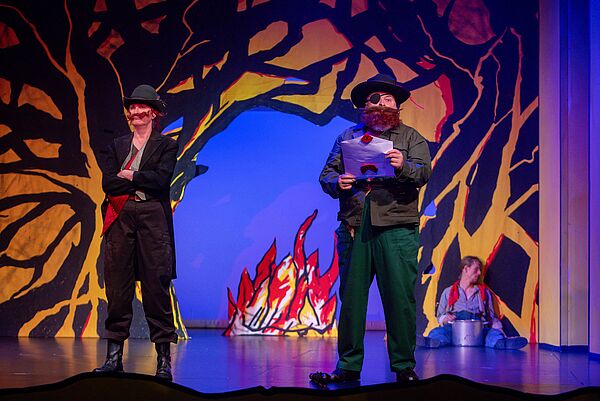 Bühnenbild. Im Hintergrund lodert ein Feuer. Zwei Räuber stehen davor, einer liest aus einem Brief.