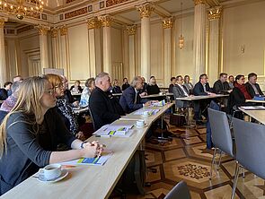 Ein historischer Saal mit Säulen und goldfarbenen Verzierungen an den Wänden. Verteilt auf drei Tischreihen sitzen rund 20 Frauen und Männer. 