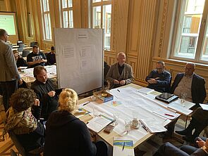 Sechs Leute sitzen an einem Tisch. Auf ihm liegen viele Zettel. Vor dem Tisch steht eine Magnettafel.