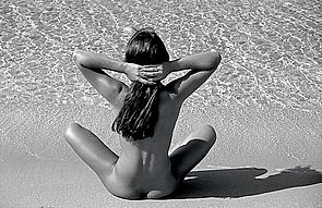 Eine nackte Frau sitzt im Schneidersitz am Strand, dort, wo Sand und Wasser aufeinandertreffen. Sie verschränkt ihre Arme im Nacken über dem langen Haar. Sie ist von hinten zu sehen.