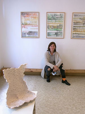Katharina Neuweg sitzt auf einem Holzbrett kurz über dem Boden. An der Wand hängen drei Bilderrahmen mit Arbeiten von ihr. Vor ihr steht eine Skulptur.