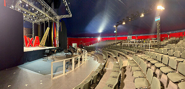 Ein Raum im Zelt. Im Raum sind eine Bühne, der Bereich für das Orchester und viele Sitzreihen.