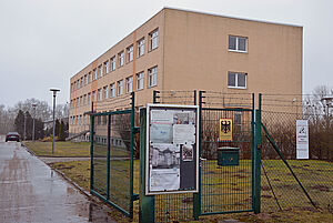 Hinter einem grünen Zaun steht ein ockerfarbenes, dreigeschossiges Gebäude. 