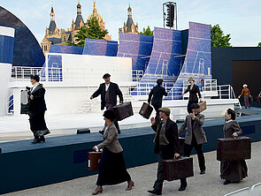 Acht Passagiere laufen mit Koffern über die Bühne.