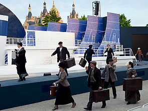 Acht Passagiere laufen mit Koffern über die Bühne.