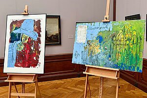 Zwei bunte Bilder auf einer Staffelei. Sie stehen in einem Raum vom Museum. Dahinter hängen Bilder an der Wand.