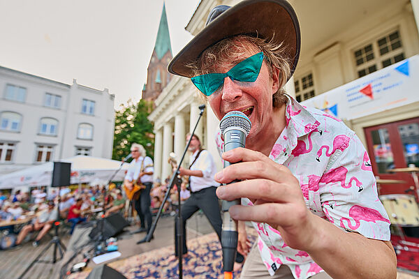 Ein Musiker singt auf einem Marktplatz. Im Hintergrund spielt seine Band.