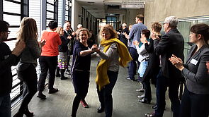 Frauen und Männer stehen auf einem Flur und tanzen und klatschen.