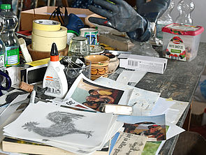 Auf einem Tisch liegen Zettel mit Zeichnungen und Bildern. Daneben stehen Flaschen und Kreppbandrollen.