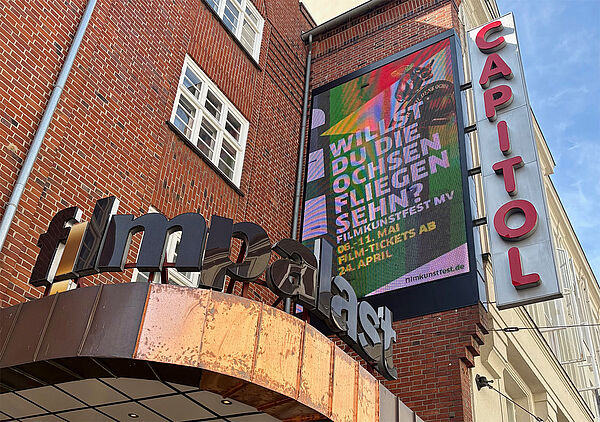 Ein Haus. An der Fassade hängt die Aufschrift Capitol. Über dem Eingang steht das Wort Filmpalast. An der Fassade hängt eine Leinwand. Sie kündigt das Filmkunstfest an. 