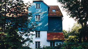 Ein vierstöckiges Haus mit blau-weißer Fassade und rotem Dach, umgeben von Bäumen.