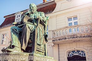 Fritz Reuter sitzt auf einem Stuhl. Eine Hand hat er ans Kinn geführt. Auf seinen Knien liegt ein Buch, das er mit einer Hand aufblättert. Das Denkmal steht auf einem Sockel. Dahinter befindet sich das Literaturmuseum. 