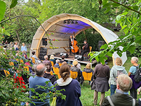 Eine Wiese. Auf der Wiese steht eine Bühne. Vor der Bühne stehen Leute. Auf der Bühne spielen Musiker.