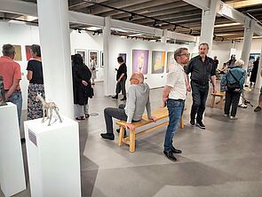 Besuchende stehen in einem Ausstellungsraum mit Bildern und Stehlen mit Skulpturen.