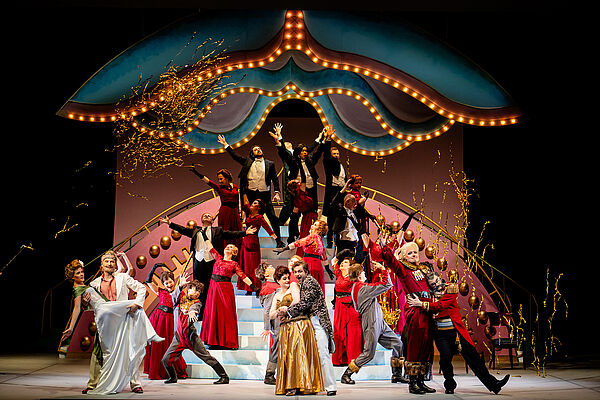 Das Bild zeigt ein opulentes Bühnenbild mit vielen tanzenden Menschen.