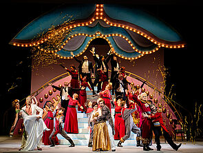 Das Bild zeigt ein opulentes Bühnenbild mit vielen tanzenden Menschen.