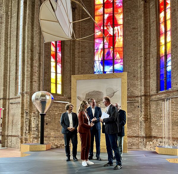 Innenraum einer Kirche mit bunten Fenstern. In der Kirche stehen vier Männer und eine Frau. Sie unterhalten sich. Ein Mann hält ein weißes Dokument in seinen Händen.