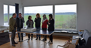 Ein Raum mit großen Fenstern. Fünf Mitglieder der Kunstkommission stehen mit dem Thomas Häntzschel an einem Tisch und betrachten Fotos. 