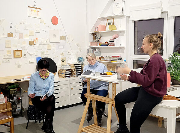 Zwei Frauen sitzen an einem Tisch in einem Atelier. Die Künstlerin sitzt auf dem Tisch und erzählt. 