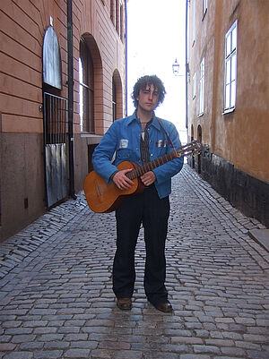 An Mann. Der Mann steht in einer engen Gasse. Er steht auf Kopfsteinpflaster. Links und rechts stehen Häuser. Der Mann hält eine Gitarre in der Hand.