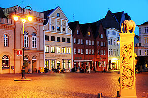 Abenddämmerung am Markt von Schwerin. Rechts: ein Denkmal mit einem Löwen auf dem Sockel. Kopfsteinpflaster ziert den Platz. Dahinter stehen Giebelhäuser. Alles ist angestrahlt.