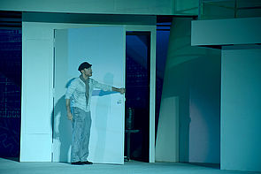 Ein Mann öffnet eine weiße Tür. Die Bühne ist in hellgrünes Licht getaucht.
