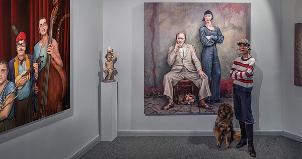 Katharina John steht in einem Raum. Neben ihr sitzt ein Hund. An den Wänden hängen große Bilder. Die Bilder stammen von Maler Manfred W. Jürgens.