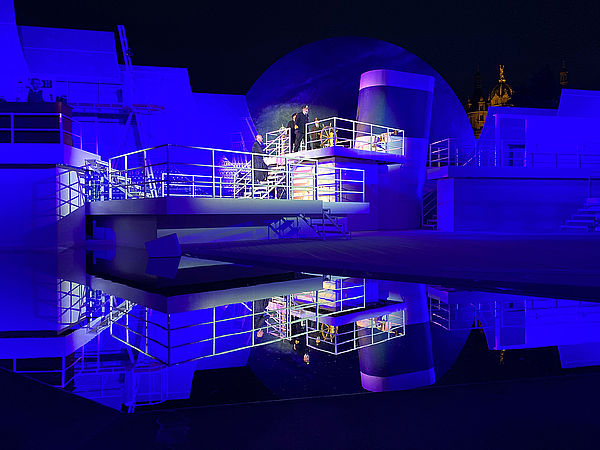 Das Bühnenbild deutet die Titanic an mit Schornstein, Brücke und Decks. Es ist blau angeleuchtet und spiegelt sich im Wasser, das vor dem Bühnenbild angelegt wurde.