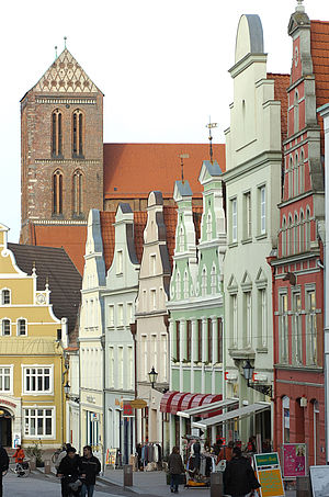 Sechs Bürgerhäuser reihen sich die Straße entlang in Richtung St. Nikolaikirche. Ihre Fassaden sind rot, weiß und grün. Vor der Kirche steht ein gelbes Bürgerhaus. Ort ist die Krämerstraße in Wismar.