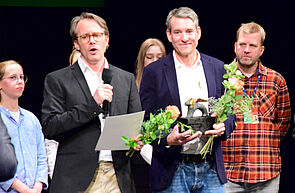 Ein Mann hält einen Preis und Blumen in der Hand. Neben ihm steht ein Mann und spricht in ein Mikrofon. Hinter ihnen stehen junge Leute. Das ist die Jury.