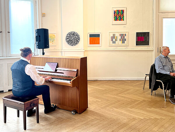 Ein Pianist spielt ein Klavier. Dahinter hängen Kunstarbeiten an einer weißen Wand in einer Ausstellung.