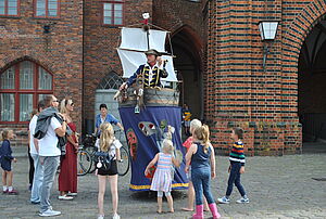 Auf dem Marktplatz in Stralsund steht ein Künstler. Passanten schauen ihm zu.