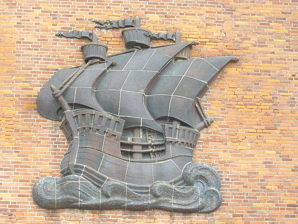 Ein Schiffswappen. Drei Segel sind gesetzt, stehen im Wind. Unter dem Schiff deutet sich Wasser an. Das Wappen befindet sich an einer Hausfassade.