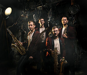 Vier Musiker. Jeder hält ein Saxofon in der Hand. Ein Scheinwerfer leuchtet auf die Musiker. Der Hintergrund liegt im Dunkeln.
