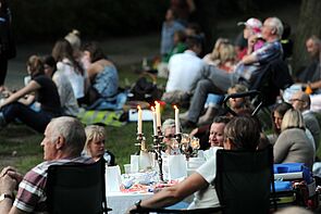 Fünf Besucherinnen und Besucher sitzen an einem gedeckten Campingtisch mit Kerzenständer. Um sie herum sitzen Menschen mit Picknickdecken auf der Wiese.