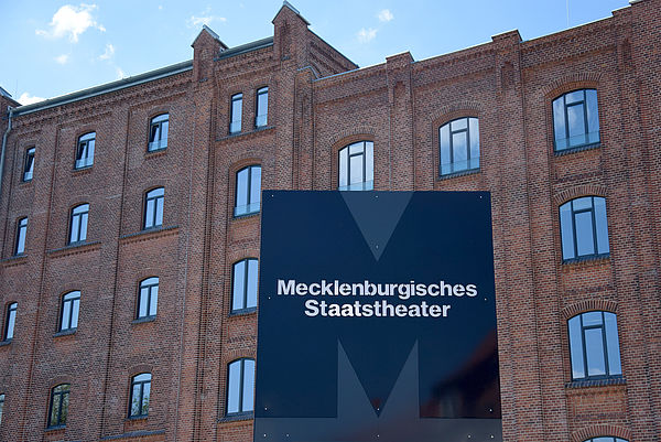 Vor dem Backsteinbau der Kulturmühle steht ein blaues Schild mit einem dunkelblauen großen M und weißer Aufschrift Mecklenburgisches Staatstheater