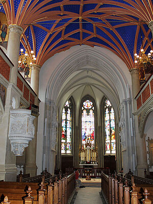 Der neugotische Chorraum mit seinem gold-blauen Deckengewölbe. Vor bunten Glasfenstern steht der Altar. Links und rechts befinden sich Sitzbankreihen. An den Seiten ragen die Emporen hervor.