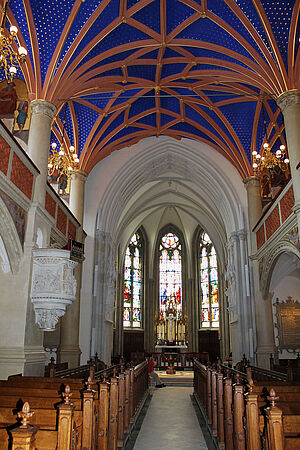 Der neugotische Chorraum mit seinem gold-blauen Deckengewölbe. Vor bunten Glasfenstern steht der Altar. Links und rechts befinden sich Sitzbankreihen. An den Seiten ragen die Emporen hervor.