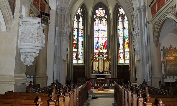Der neugotische Chorraum mit seinem gold-blauen Deckengewölbe. Vor bunten Glasfenstern steht der Altar. Links und rechts befinden sich Sitzbankreihen. An den Seiten ragen die Emporen hervor.