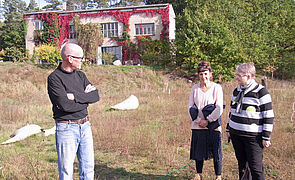 Drei Personen stehen auf einer Wiese. Im Hintergrund steht ein Haus. 