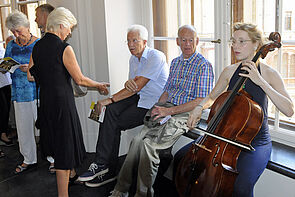 Eine Frau spielt vor dem Plenarsaal Cello. Direkt neben ihr sitzen und stehen Zuschauerinnen und Zuschauer.