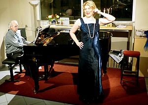 Britta Kopelke steht in einem blauen Abendkleid vor einem Klavier. Reinhard Lippert sitzt am Klavier und spielt.