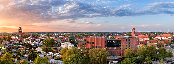 Ein Luftbild von Parchim. Im Vordergrund steht die Kulturmühle. Sie ist ein altes Backsteingebäude. Im Hintergrund stehen weitere Häuser der Stadt. Aus der Silhouette ragen zwei Kirchen hervor. 