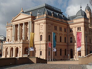 Das Schweriner Theater von außen. Es trägt eine ockerfarbene Fassade. Der Eingang führt durch einen kleinen Vorbau, der ein Stockwerk höher als Balkon dient.