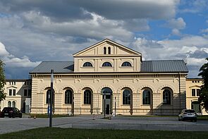 Außenaufnahme vom Marstall. Das Gebäude trägt eine beige Fassade. Links und rechts befinden sich drei halbbaumgroße Fenster, dazwischen die Eingangstür. Dunkle Wolken mischen sich in den blauen Himmel.